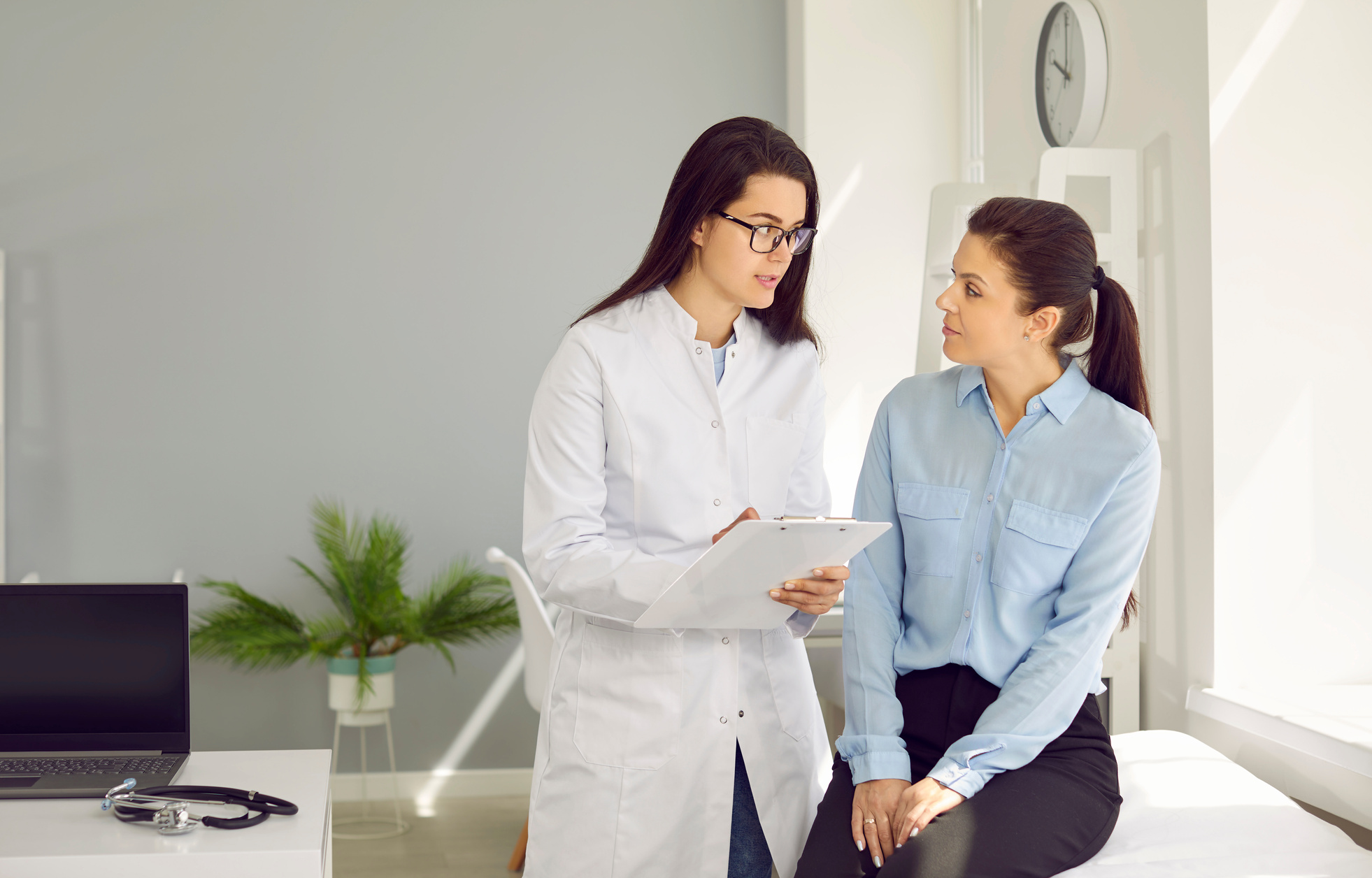 Young Woman Doctor in White Medical Coat Consults Female Patient Sits on Hospital Couch